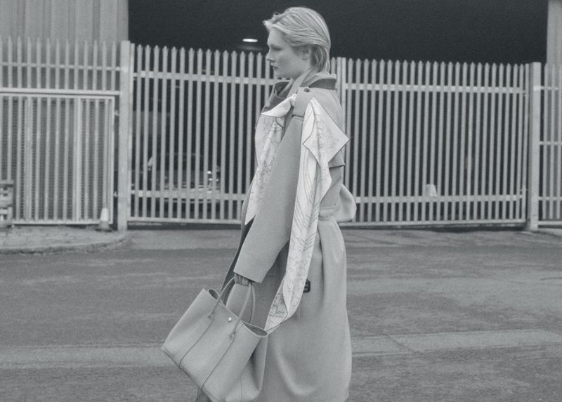 Monochrome fashion shot: A woman in a trench coat carries a tote bag in an urban setting with metal fence in the background.