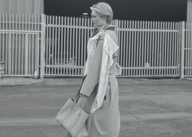Monochrome fashion shot: A woman in a trench coat carries a tote bag in an urban setting with metal fence in the background.