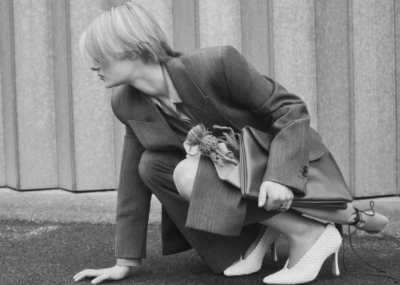 A person in a suit and heels crouches on asphalt, holding a purse, in a black and white fashion photograph with a textured background.