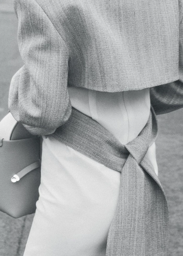 Close-up of a person wearing a grey cropped jacket with a tied wrap belt and holding a structured handbag.