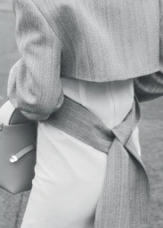 Close-up of a person wearing a grey cropped jacket with a tied wrap belt and holding a structured handbag.