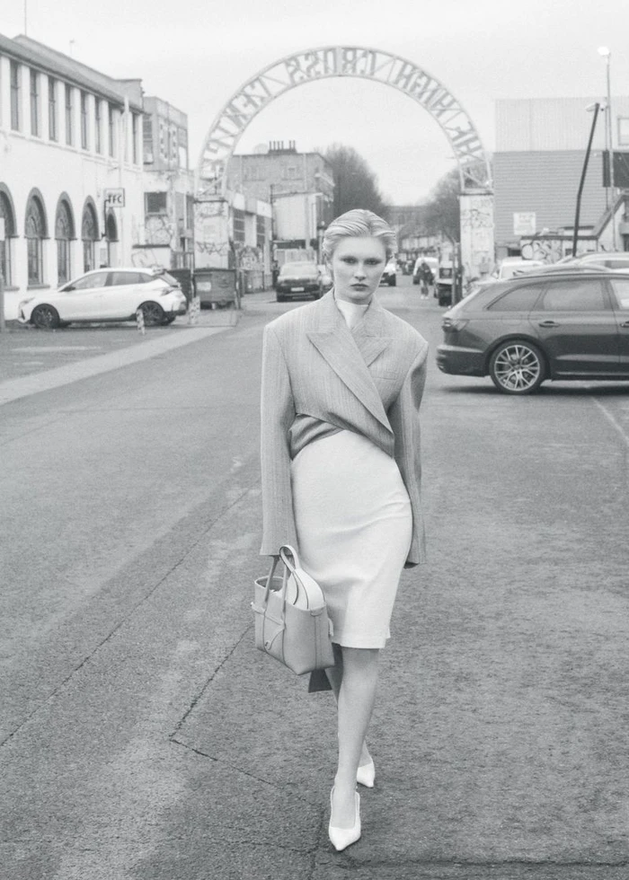 Model walking down a paved street carrying a handbag wearing a structured blazer and dress