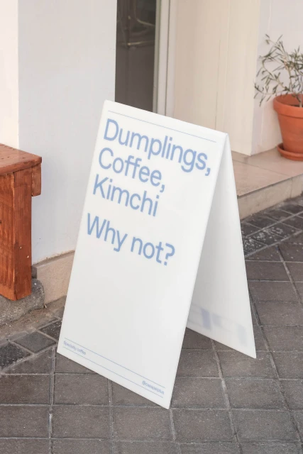 A-frame sign outside a cafe advertising dumplings, coffee, and kimchi with the playful invitation, 'Why not?' stands on a paved sidewalk.