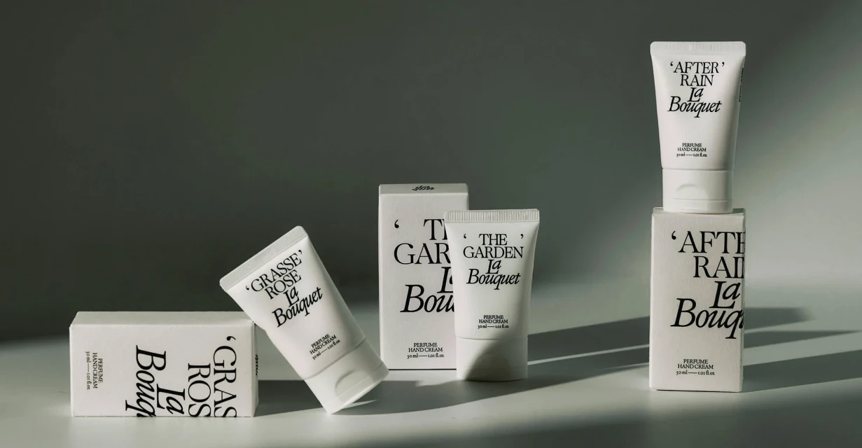 Product shot of 'La Bouquet' hand cream tubes and boxes in neutral tones, arranged on a light surface against a grey gradient background.