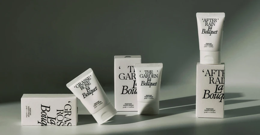 Product shot of 'La Bouquet' hand cream tubes and boxes in neutral tones, arranged on a light surface against a grey gradient background.