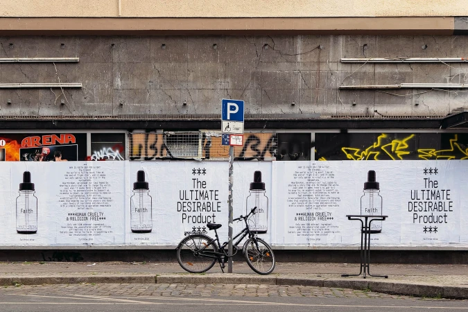Urban scene with a bicycle parked in front of posters advertising a product called Faith, emphasizing human cruelty and religion-free values.