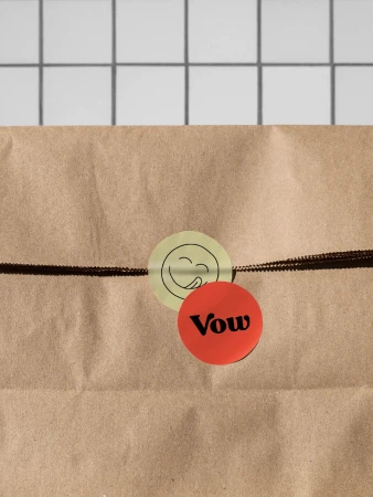 A brown paper bag is sealed with a round yellow sticker featuring a simple cartoon face and a red sticker with the word 'Vow' on it.