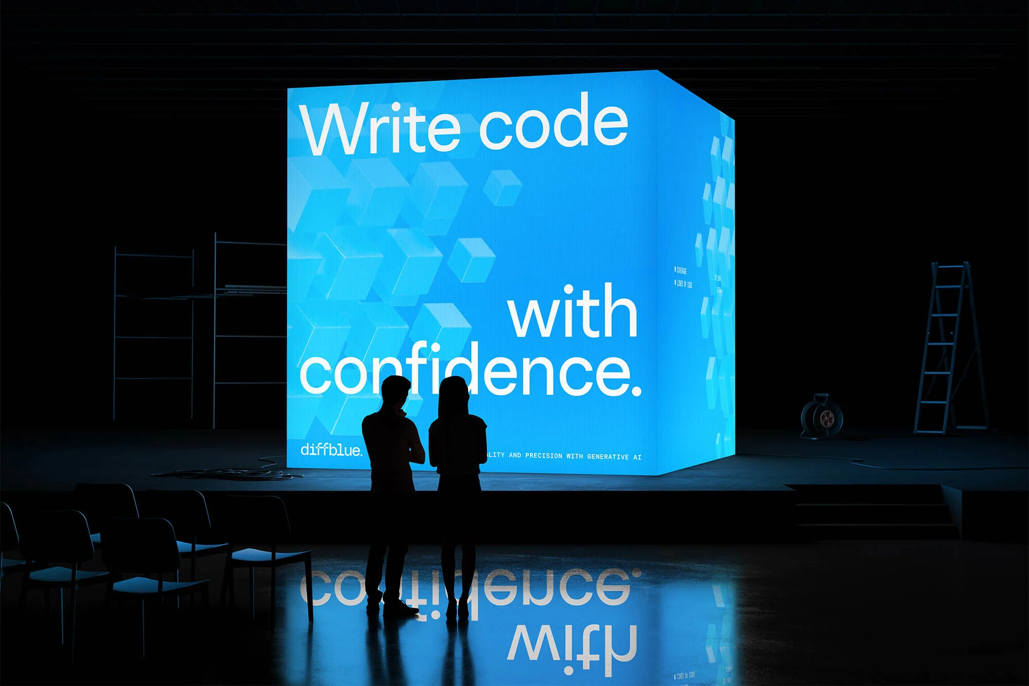 Two people look at a large blue cube on a stage with the text 'Write code with confidence.' Chairs and a ladder are visible in the scene.