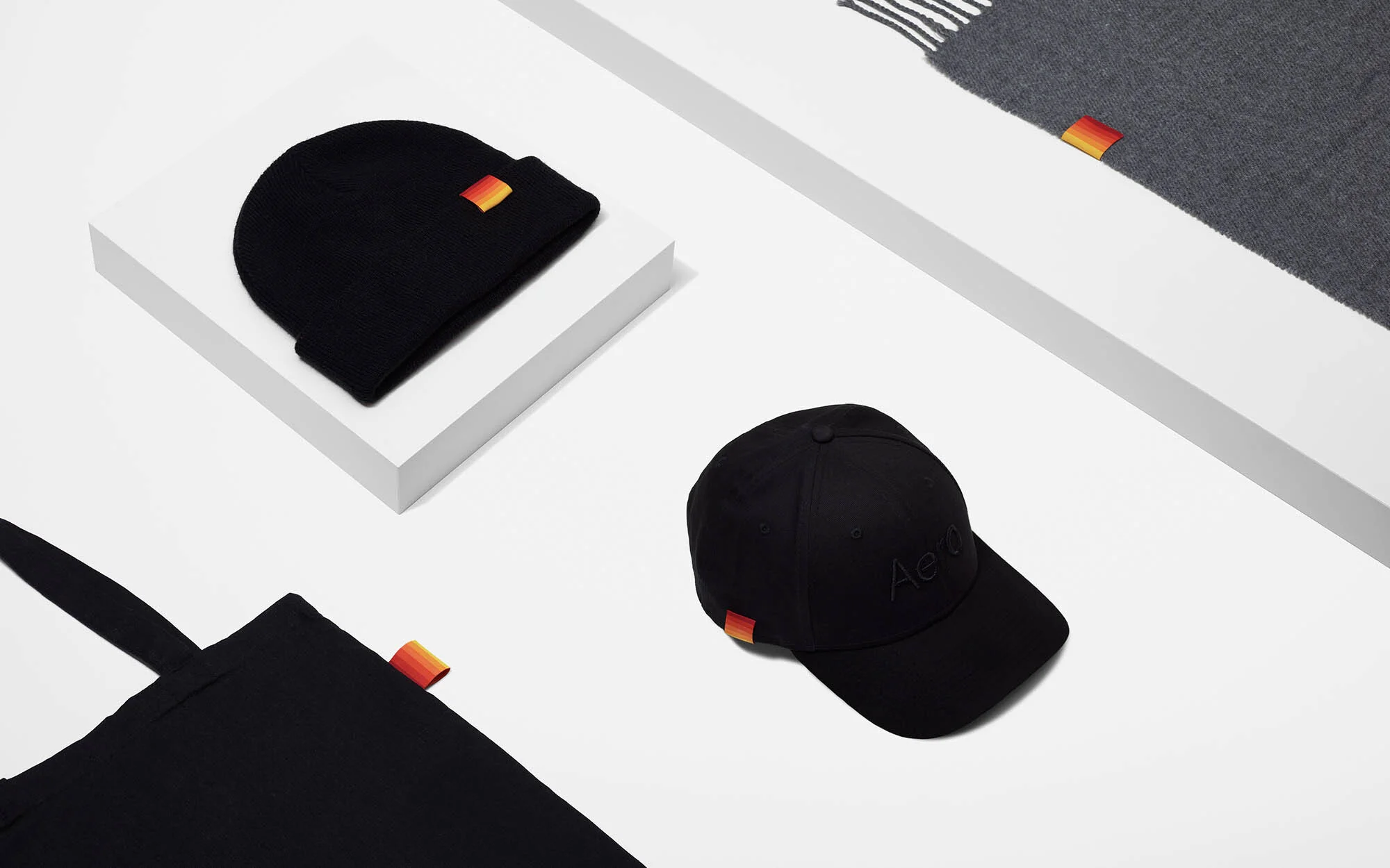 Flat lay of black beanie, baseball cap, tote bag, and gray scarf on a white platform. Product photography featuring fashion accessories.