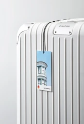 White ridged suitcase with a metallic corner plate and hanging luggage tag showing a building exterior against a white background
