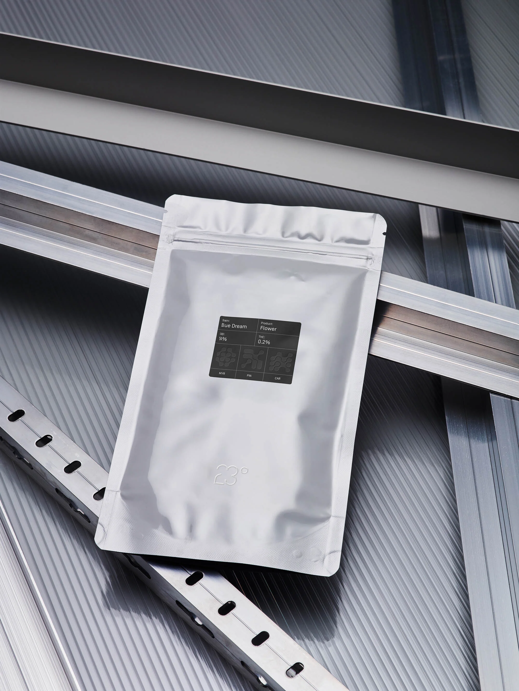Silver, sealed package with a black label sits diagonally on a ridged, metallic surface with open metal framing in the background.