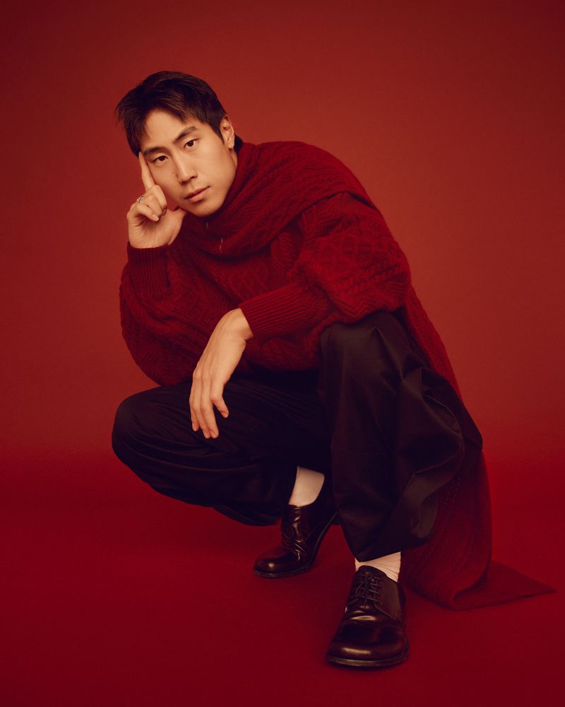 Portrait of a young Asian man crouching against a red background, wearing a red sweater, black pants, and black dress shoes for a fashion shoot.