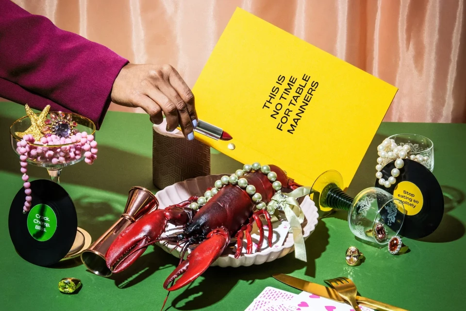 Still life composition with a lobster wearing a pearl necklace on a green surface, surrounded by jewelry, records, and a yellow sign.