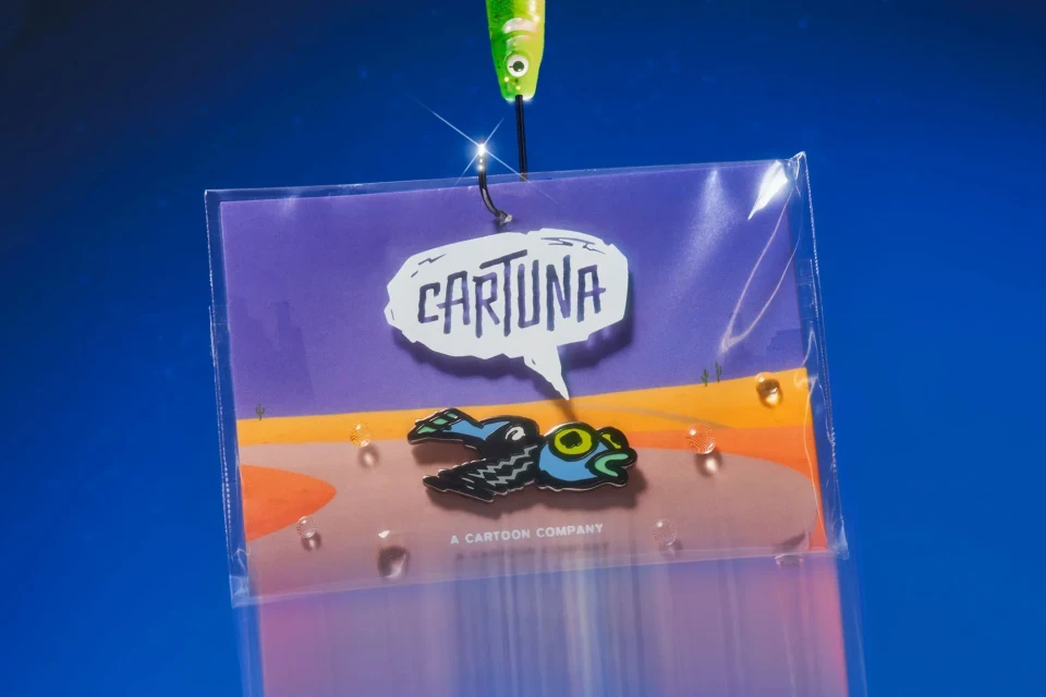 Fishing lure presentation of colorful cartoon fish-shaped pin on card, against gradient blue background, commercial product shot.