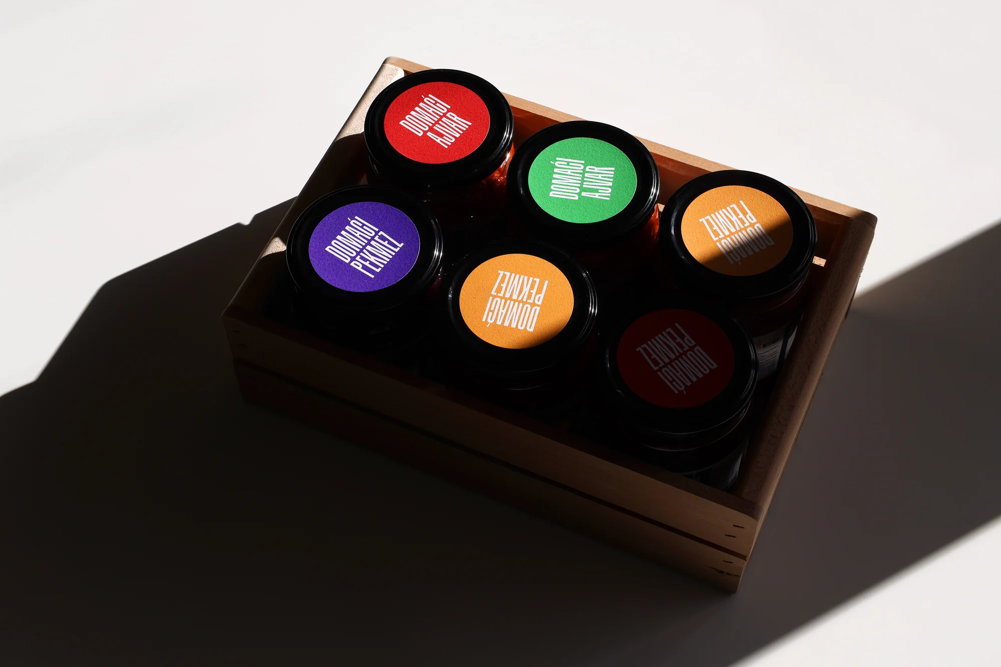 Overhead shot of six glass jars with colorful labels in a wooden box, casting shadows on a white surface, studio lighting.