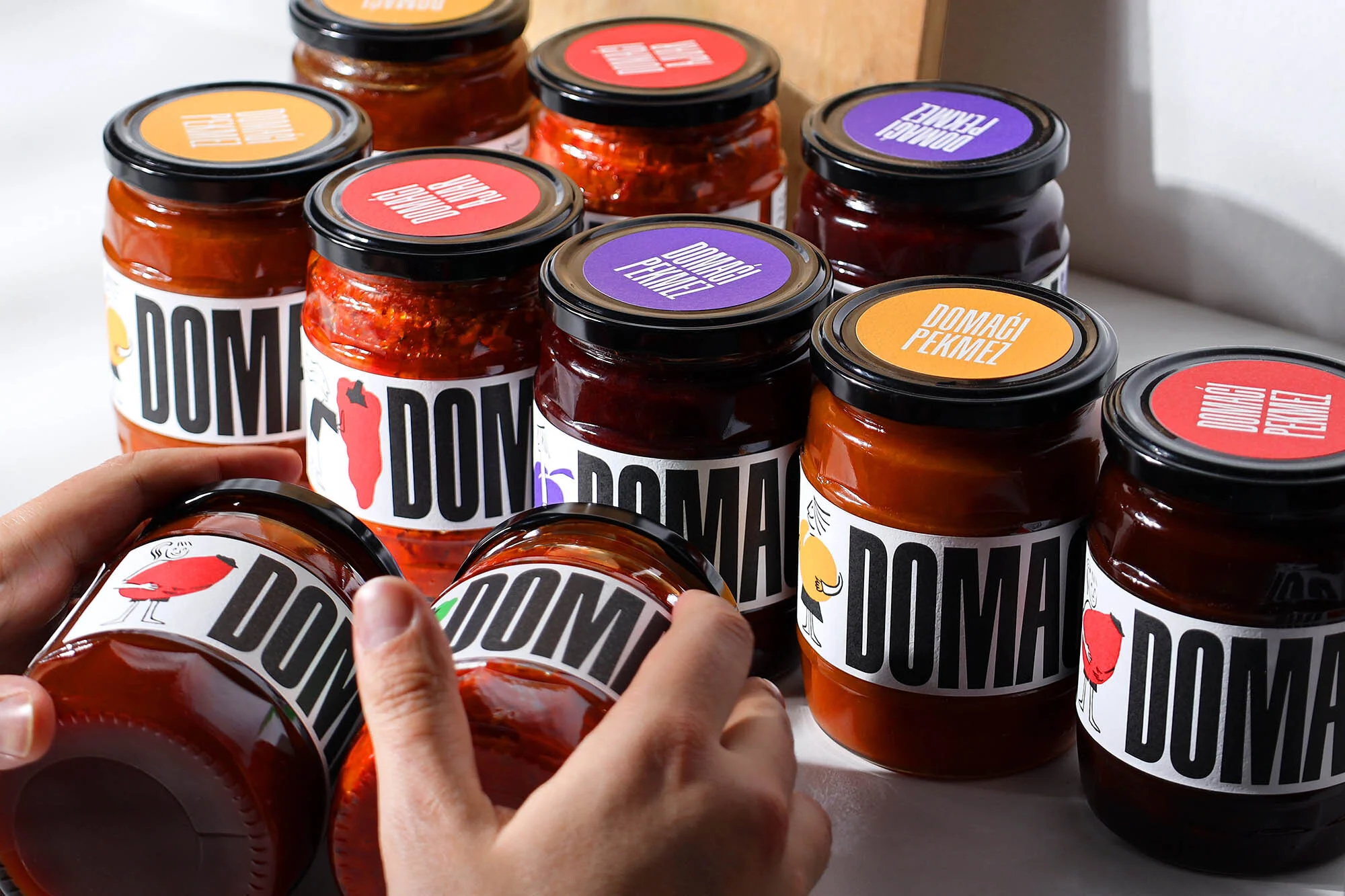 Various flavors of DOMAGI PEKMEZ fruit preserves in glass jars with colorful lids, some being held in human hands, on a white surface.