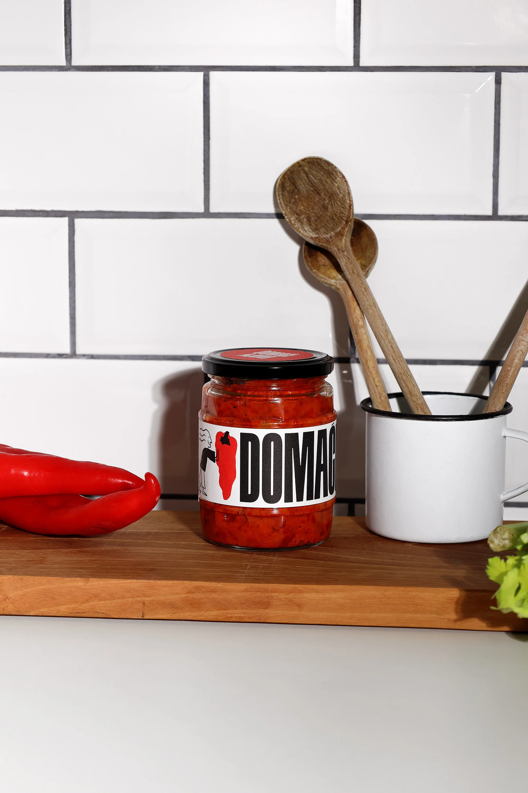 A jar of red spread sits on a wooden board next to red peppers and wooden spoons with a white tile background. Food and beverage.
