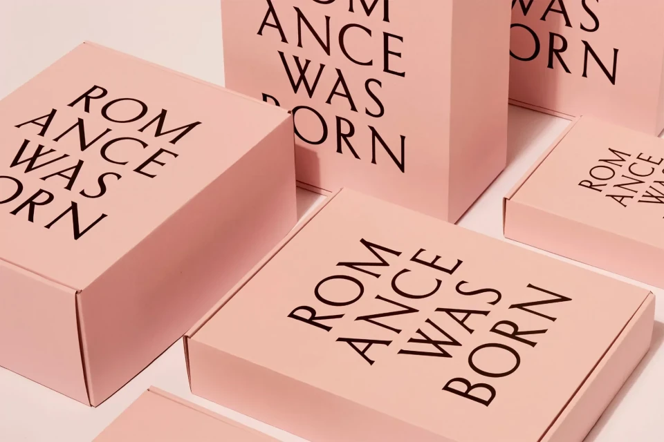 Several light pink rectangular boxes and one square box are stacked with the text ROMANCE WAS BORN in black block letters, product shot.