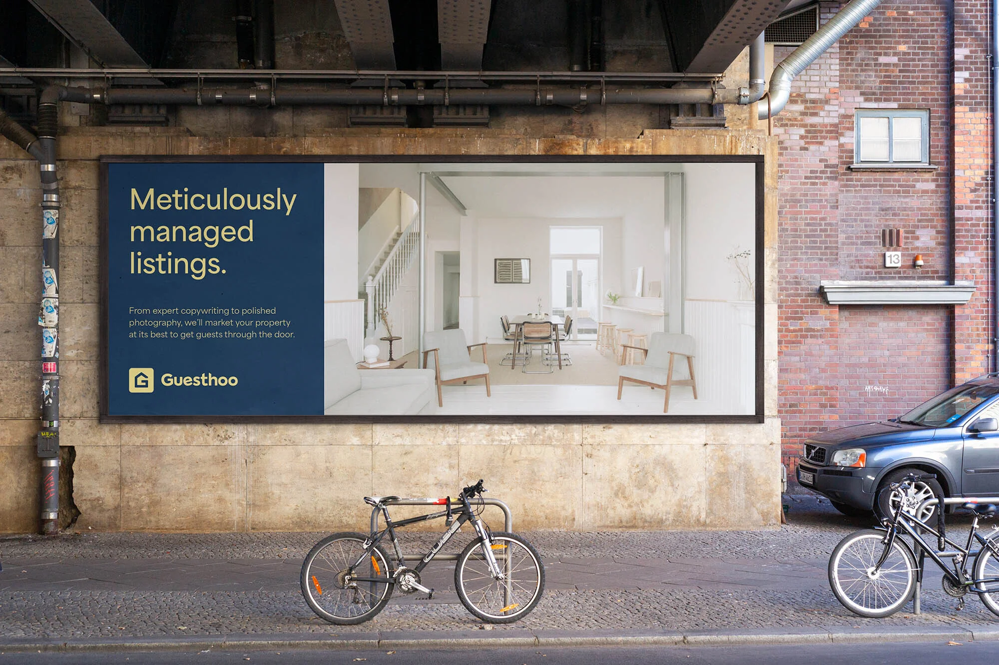 Billboard advertisement for Guesthoo featuring interior design, mounted on a building, with bicycles and a car parked in the foreground.
