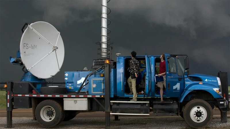 Blue First Street weather tracking truck with satellite dish and two people standing near the open doors, against a dark stormy sky.