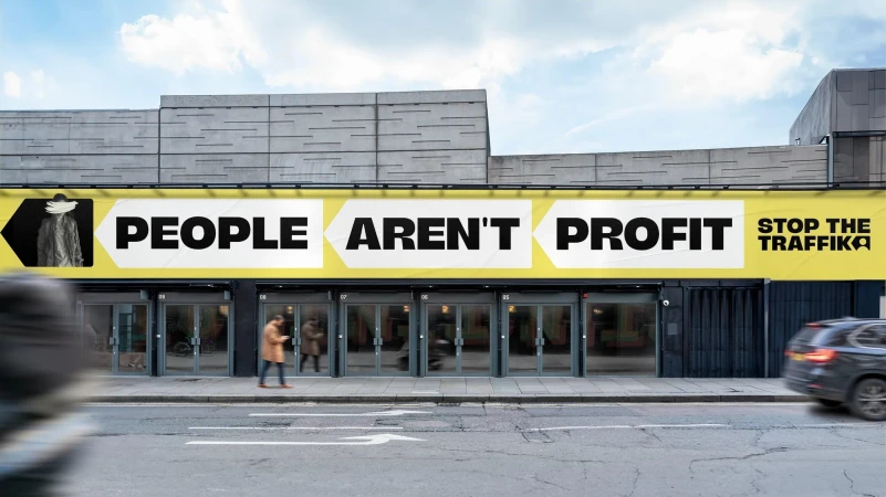 Building facade displays 'People Aren't Profit' banner, with people walking nearby and a car passing on the street.