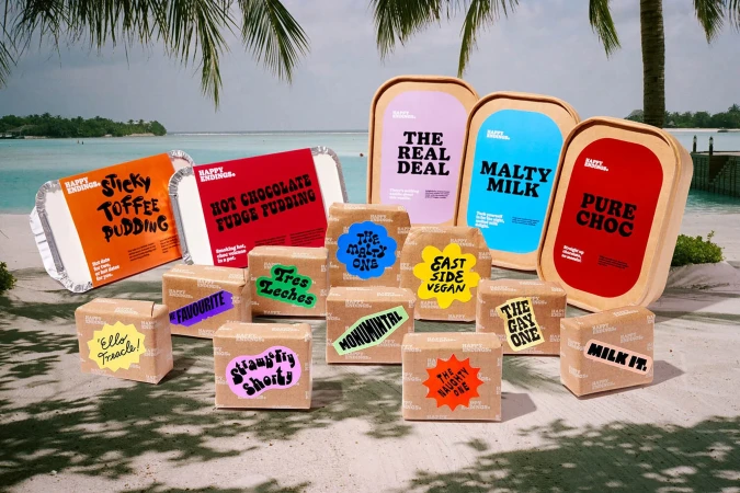 Selection of Happy Endings dessert boxes and trays arranged on a beach with blue ocean and palm trees in the background. Colorful packaging.