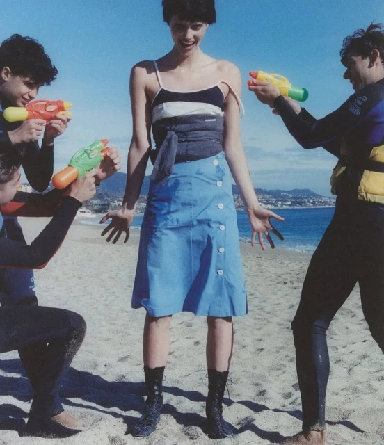 Fashion shot of a female model in a skirt, with two men on either side pointing water guns at her, set on a beach with sea views.
