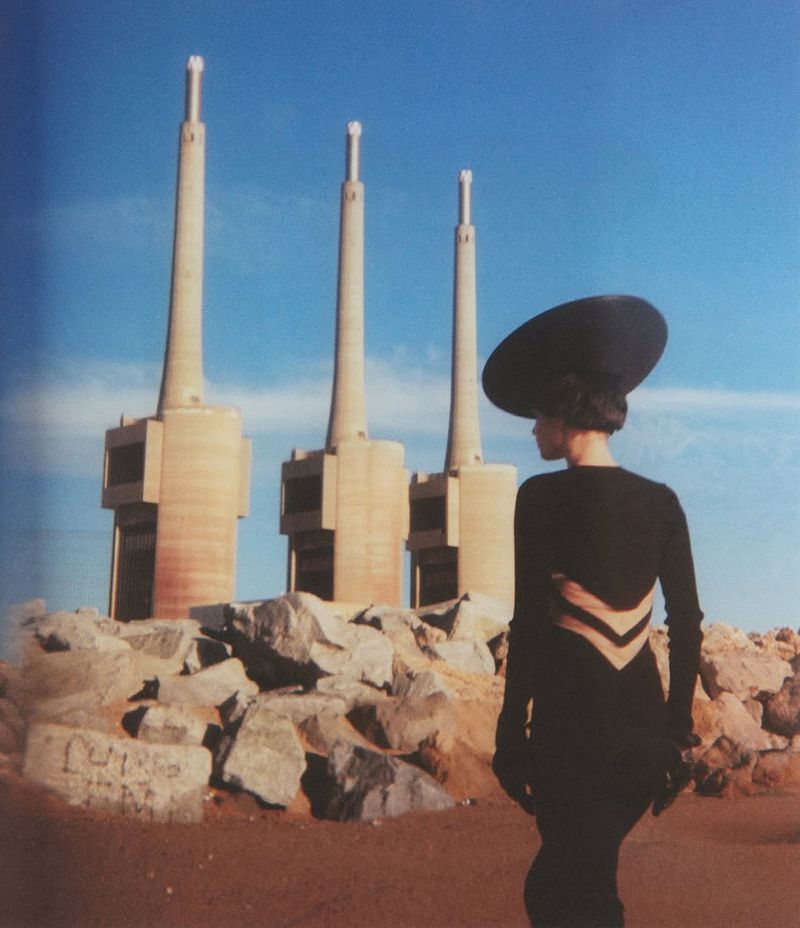 Adult female fashion model in black dress and hat posing in rocky desert landscape with industrial towers under bright blue sky.