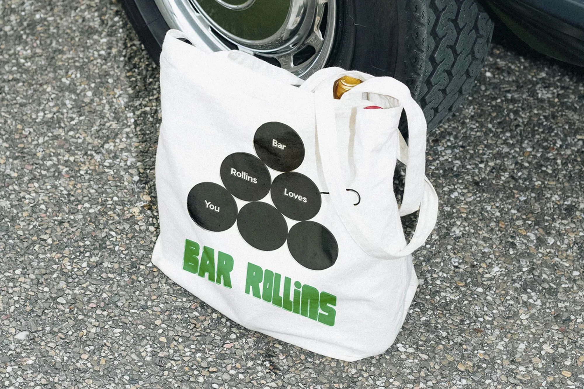 Tote bag with 'Bar Rollins' printed in green next to a car tire on asphalt. The tote bag features graphics with circles.
