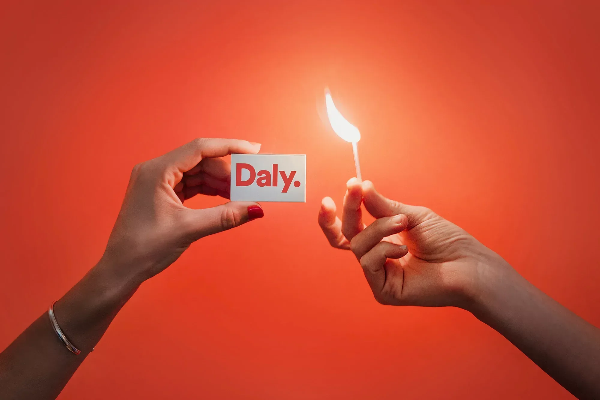Woman's hands holding matchbox with Daly logo and lit match on a vibrant orange background, simple and striking composition.