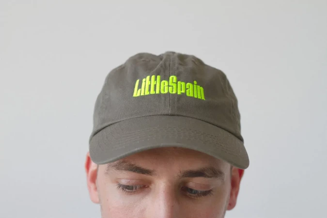 Close-up of a person wearing a grey baseball cap with the neon green text 'LittleSpain' embroidered on it against a white background.