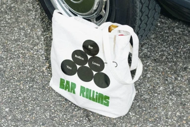 White canvas tote bag with black circular graphic and green text resting on asphalt pavement next to car tire