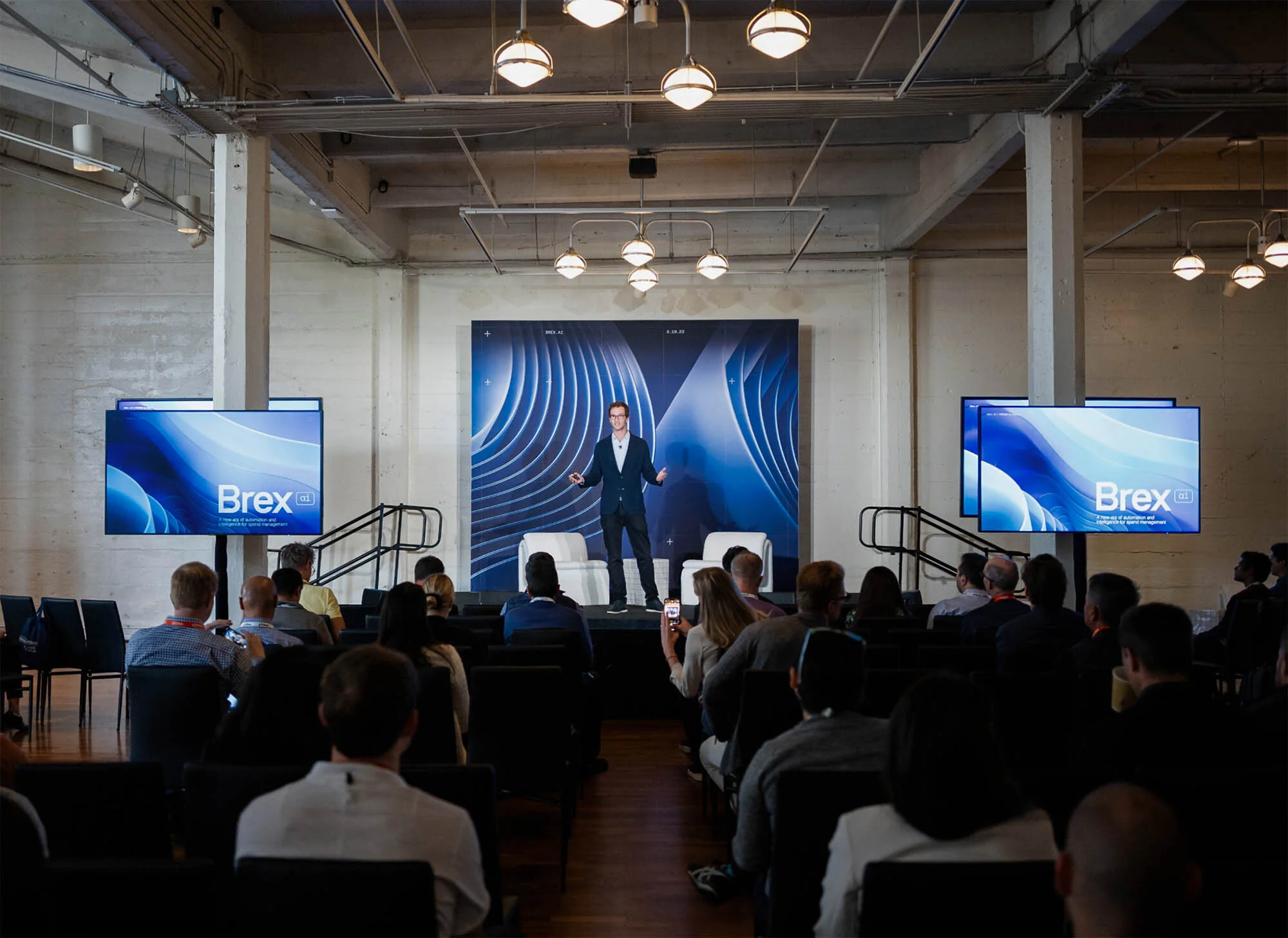 Corporate event featuring a presenter on stage in front of a screen, addressing an audience seated in rows in a large event space.