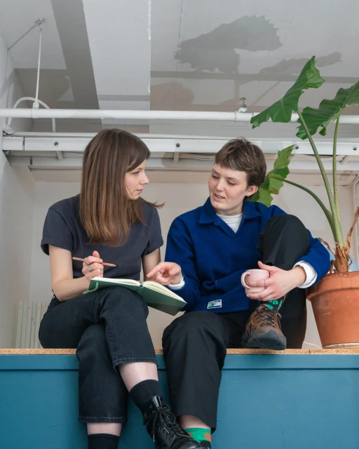 Two women sitting on a ledge, discussing a notebook in a brightly lit interior space with a large potted plant in the background.