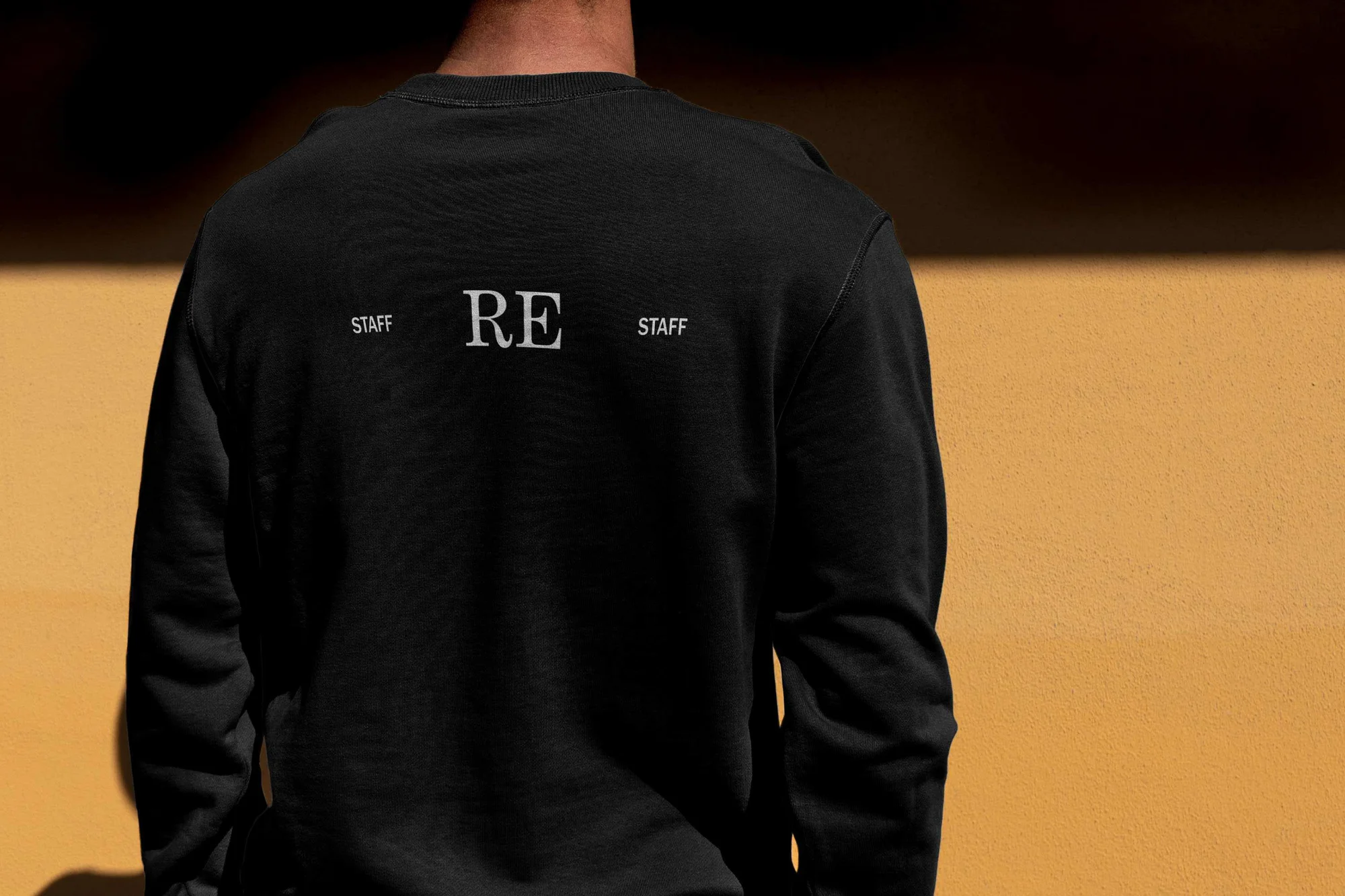 Back view of a person wearing a black long-sleeve sweater with 'RE STAFF' text, standing against a textured tan color wall.