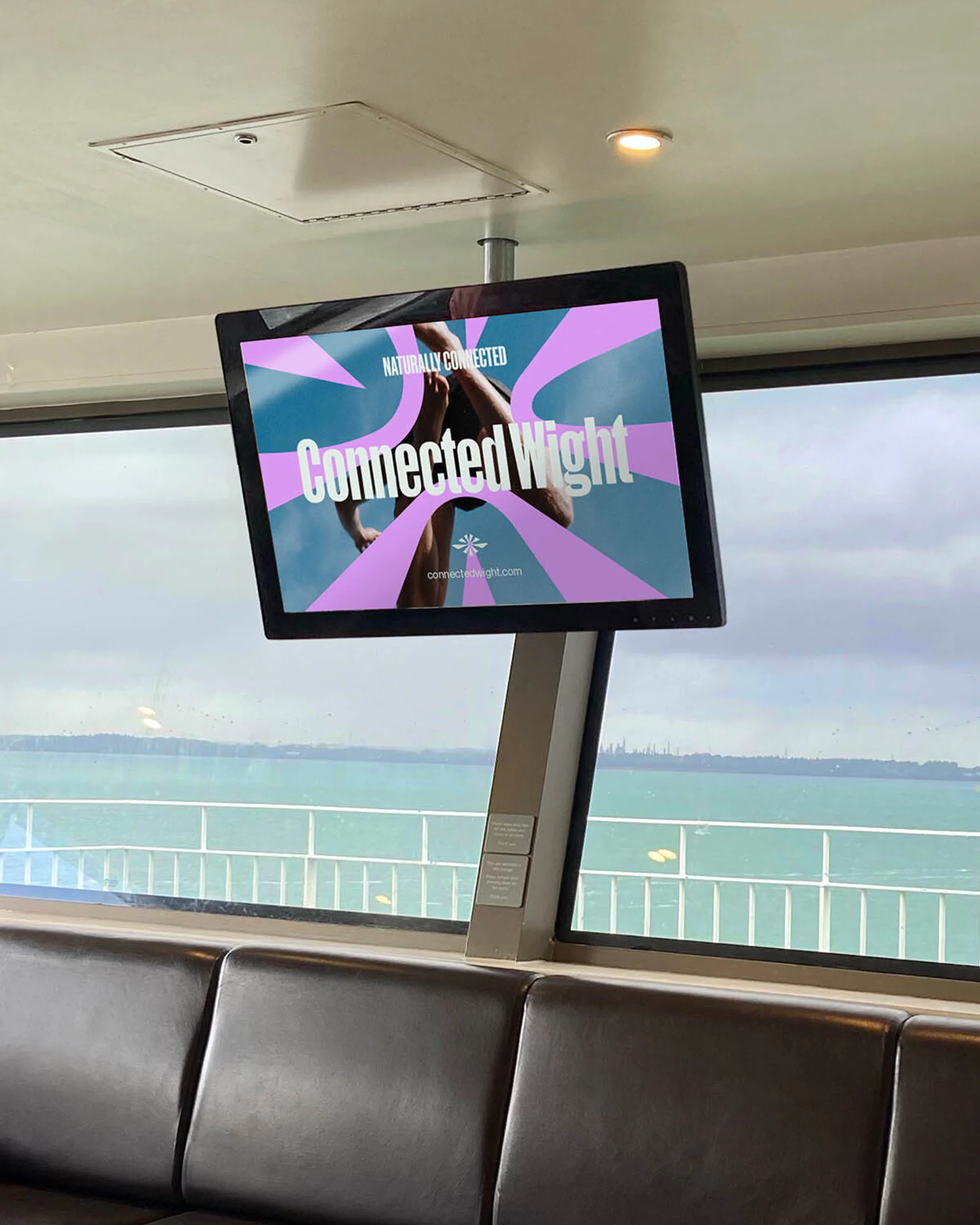 Indoor view featuring an advertisement screen on a ferry, showcasing a fitness promotion, along with visible seating and a window to the sea.