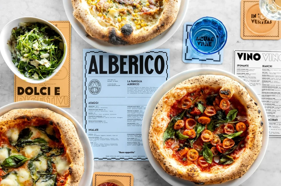 Overhead shot of a table with three pizzas on plates, a bowl of salad, drinks, and restaurant menus, showcasing a food and beverage theme.