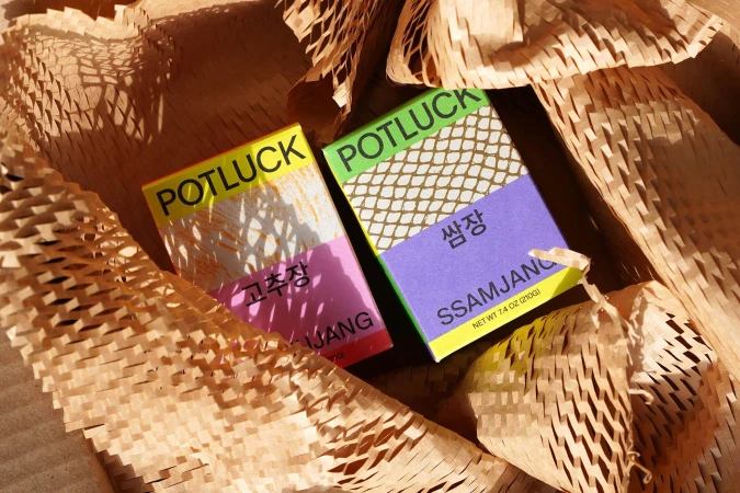 Two Potluck brand Gochujang and Ssamjang sauce packets nestled in tan packing material, casting shadows; food and beverage packaging.