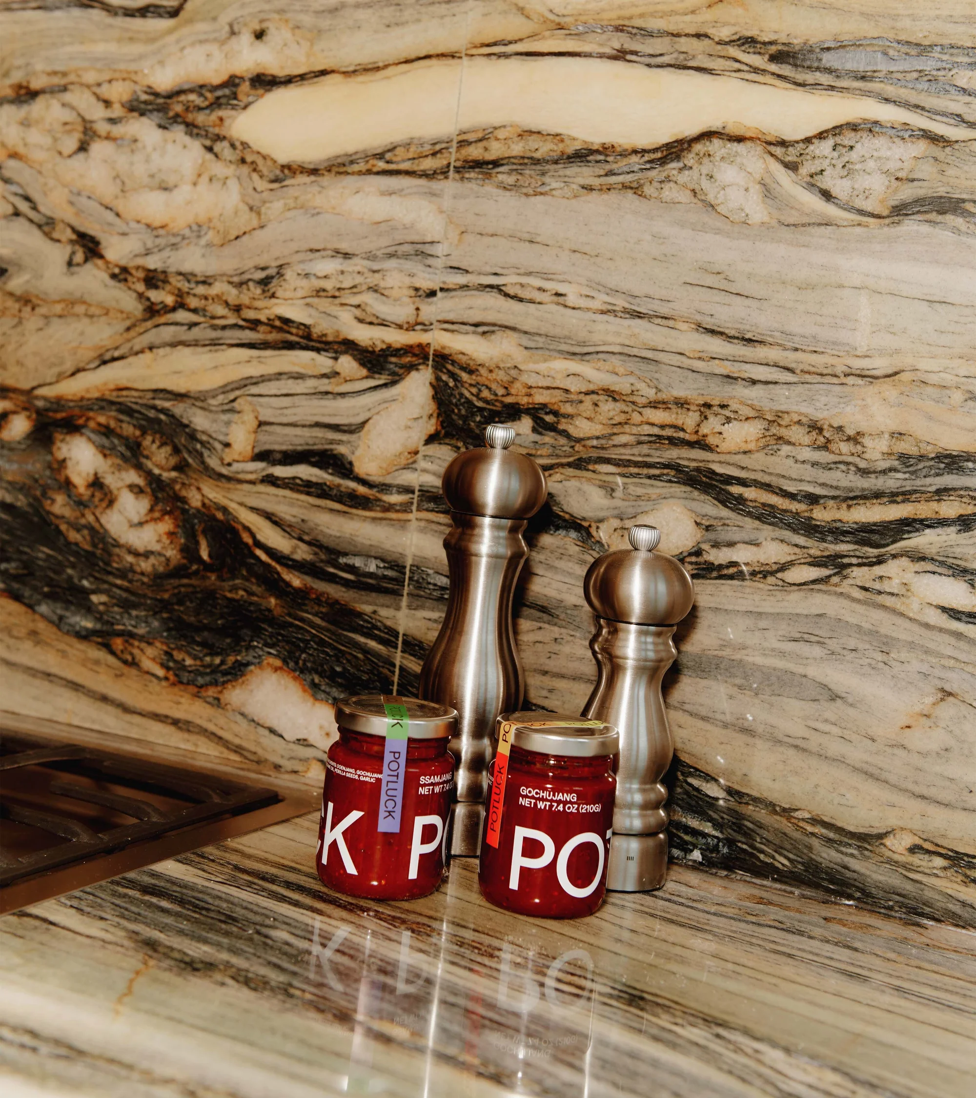 Close-up of two jars labeled 'Potluck' beside two metal pepper mills on a marble countertop with a dark marble backsplash.