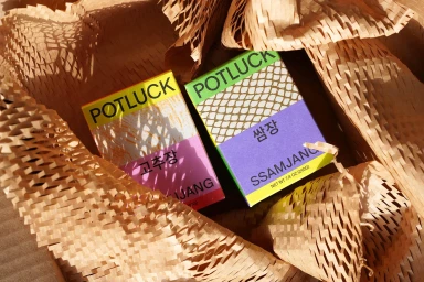 Two colourful Potluck brand product boxes nestled inside crinkled brown packing paper.
