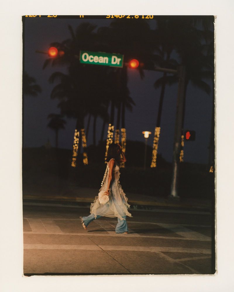 A woman in a fashionable dress walks across a crosswalk at night, with palm trees and city lights in the backdrop under red traffic lights.