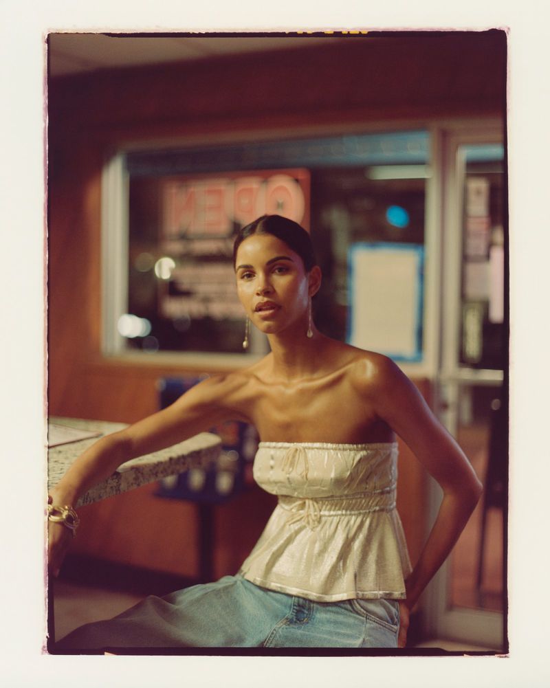Young woman poses in a vintage-style diner at night, wearing a strapless top and jeans, exuding a casual yet elegant style.