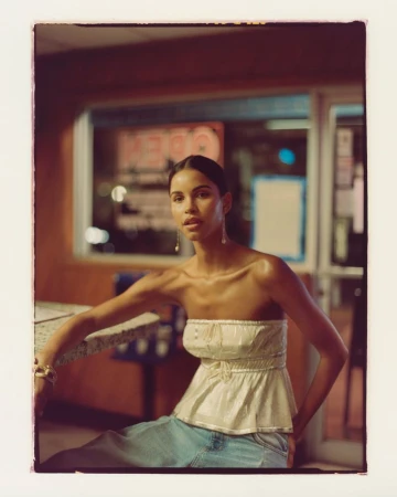 Young woman poses in a vintage-style diner at night, wearing a strapless top and jeans, exuding a casual yet elegant style.
