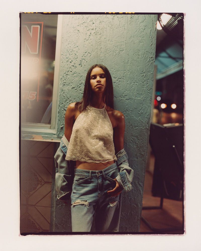 A young woman with long hair poses in a halter top and jeans, leaning against a textured wall with neon light reflecting in the background.