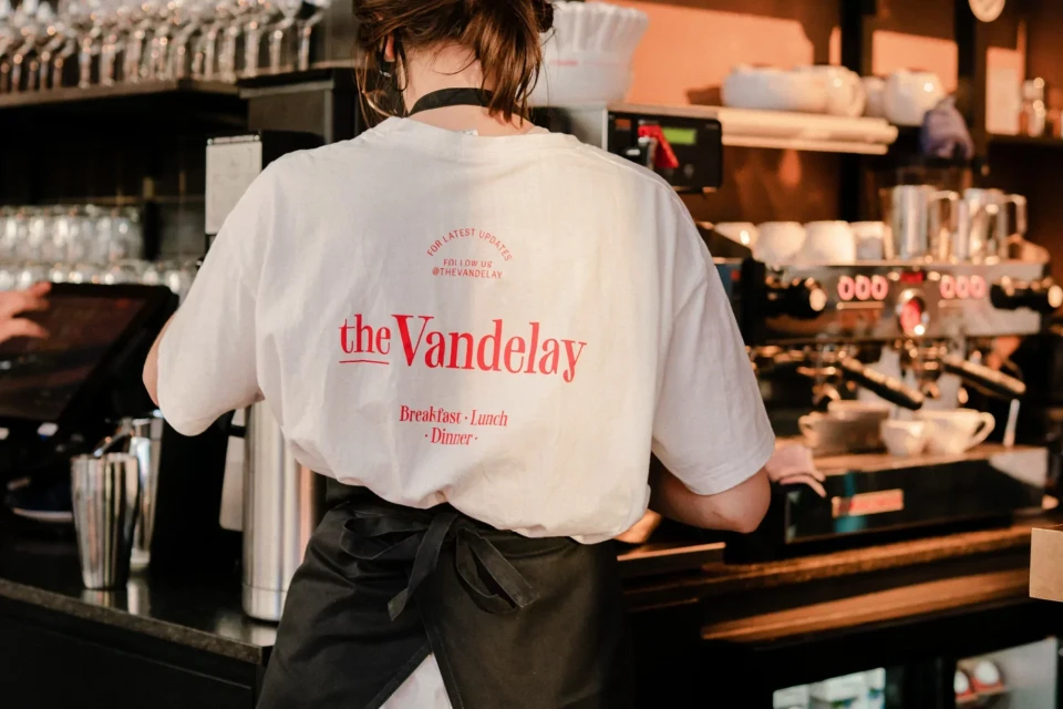 A person stands at a cafe espresso machine wearing a Vandaleay shirt. Bar and glassware visible. Serving up coffee.