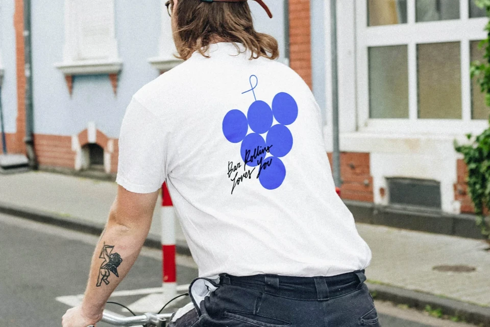 Man on bicycle in white tee shirt with blue graphic design against a street scene background, seen from the back.