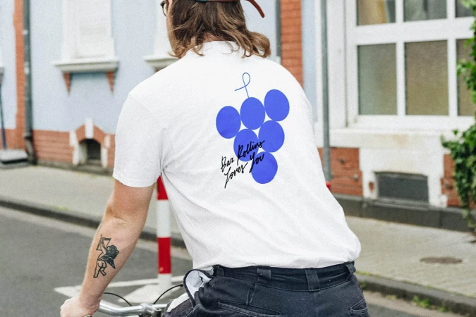 Man on bicycle in white tee shirt with blue graphic design against a street scene background, seen from the back.