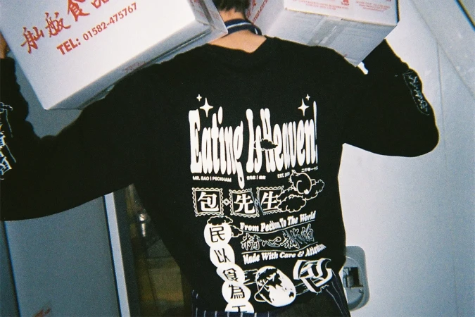 Person wearing a black long-sleeved shirt with text and graphic designs while carrying boxes in an indoor setting with soft lighting.