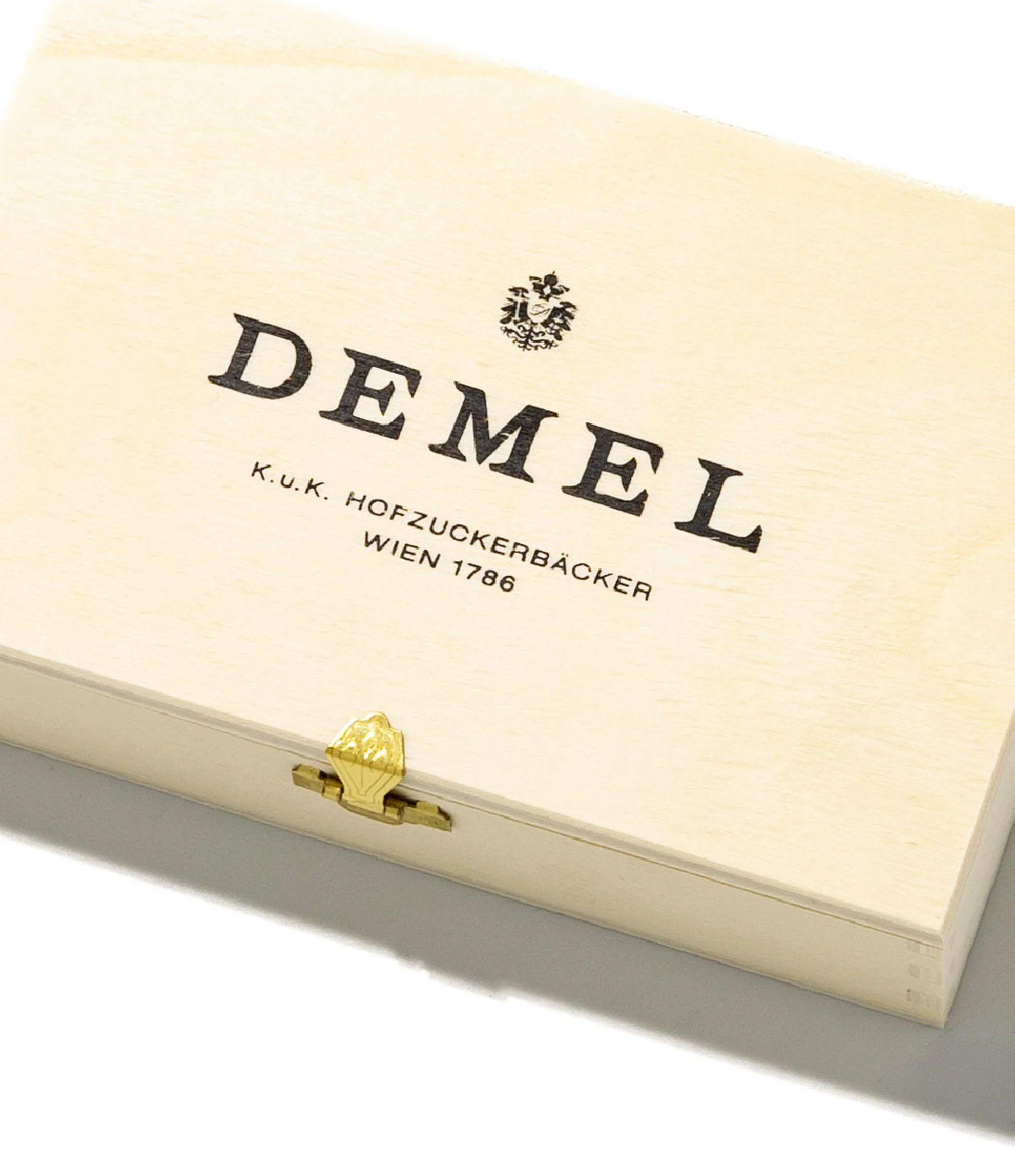 Close-up of a light-colored wooden box with a gold latch, featuring the Demel logo and brand information printed in black ink.