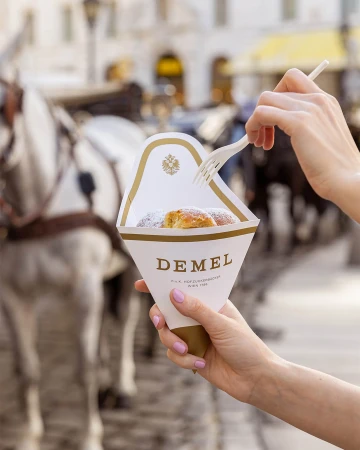 Woman enjoys a Demel pastry outdoors, holding a paper cone filled with the treat and using a fork, with blurred horses in background.